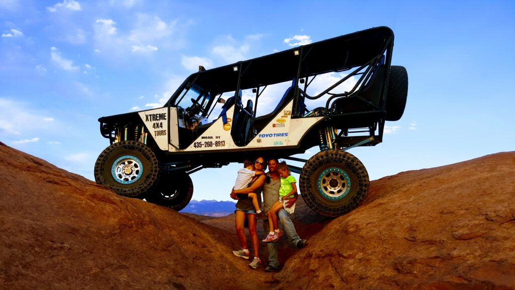 family posing beneath open-air 4x4 vehicle on Moab off-road trail